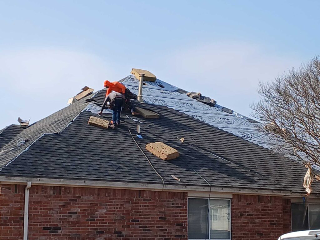 Two roofers are actively working on a residential roof, with one section covered in blue underlayment and another section already shingled with dark asphalt shingles.