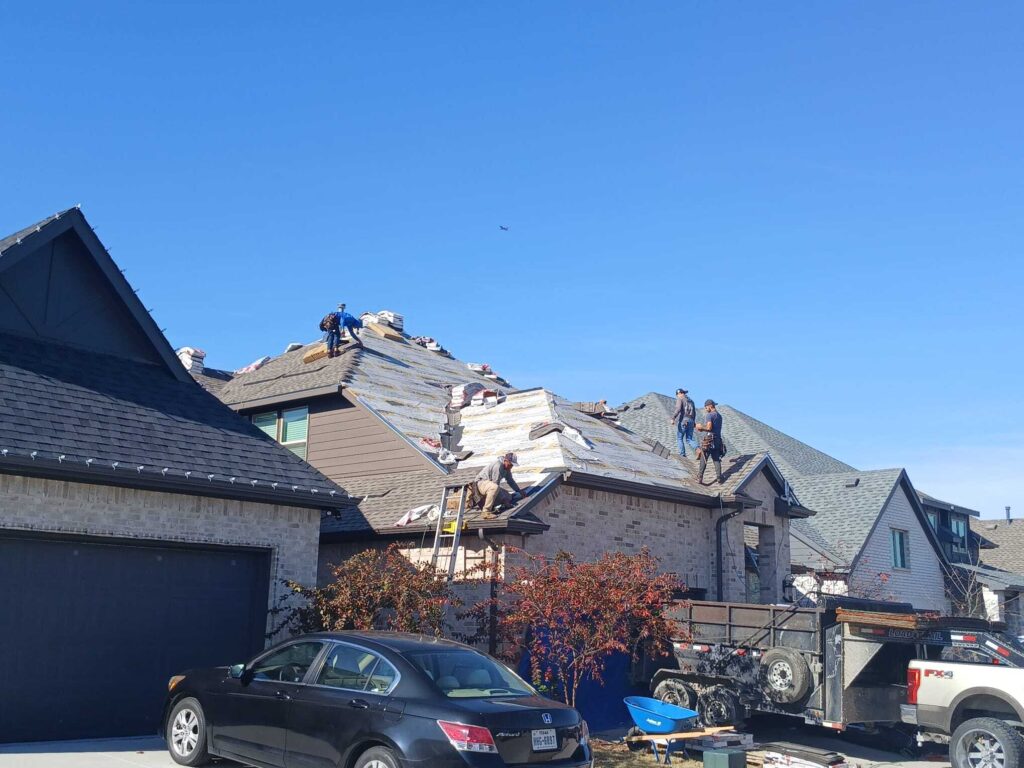 Several roofers are actively working on a residential roof, which is partially stripped down to the underlayment, with bundles of new shingles visible. A dump trailer and a work truck are parked in the driveway, and a black sedan is also visible.