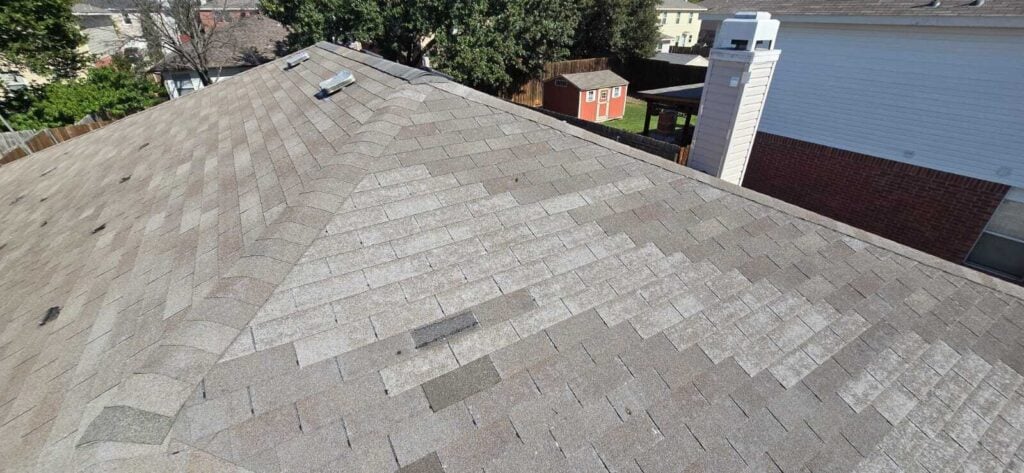 An aerial view of an old, worn asphalt shingle roof showing significant granule loss, discoloration, and some missing shingles. The surrounding neighborhood, including a red shed and a chimney, is visible in the background.