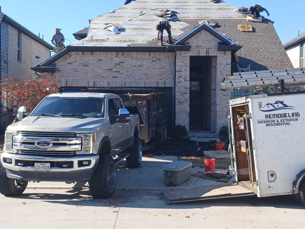 This image shows a residential house with several roofers actively working on the roof, which is partially stripped down to the underlayment and partially being re-shingled. A Ford Super Duty truck and a utility trailer are parked in the driveway.