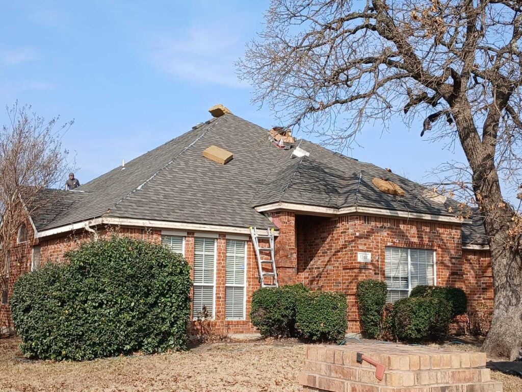 A brick house with an asphalt shingle roof undergoing replacement, with a worker visible on the roof and bundles of shingles placed on the slopes. A ladder leans against the front of the house.