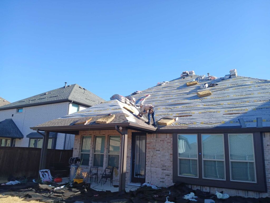 A residential house is undergoing roof replacement, with one worker visible on the roof installing new asphalt shingles over the underlayment. Debris from the old roof is scattered on tarps on the ground below.
