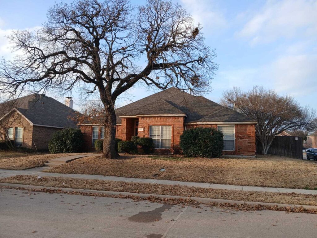 A single-story brick house with an asphalt shingle roof is visible from the street, featuring a large, bare tree in the front yard and dormant grass.