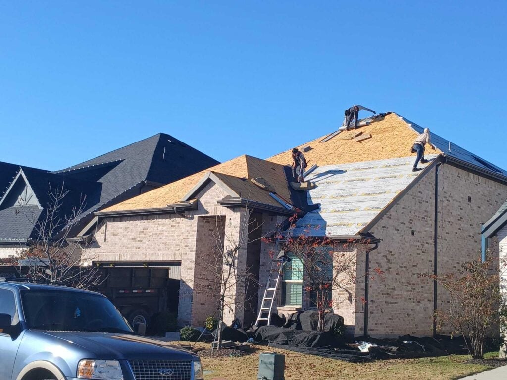 Several roofers are actively working on a residential roof, with sections showing exposed decking, underlayment, and new roofing material being installed. A dump trailer is parked in the driveway, and protective tarps are laid around the house.
