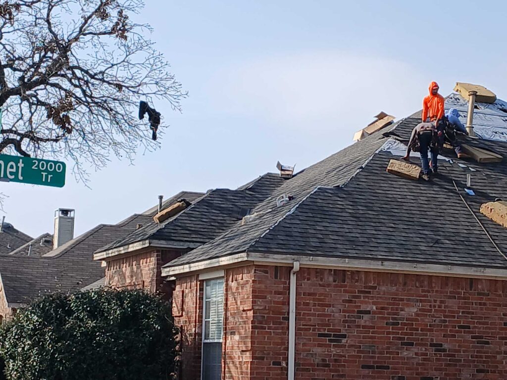 Several roofers are actively working on a residential roof, installing new asphalt shingles over visible underlayment. Boxes of new shingles are scattered on the roof, and a street sign is visible in the upper left.