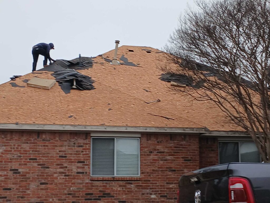 A worker is actively stripping old asphalt shingles from a residential roof, exposing the plywood decking. The brick house and a partial view of a dark truck are visible below.