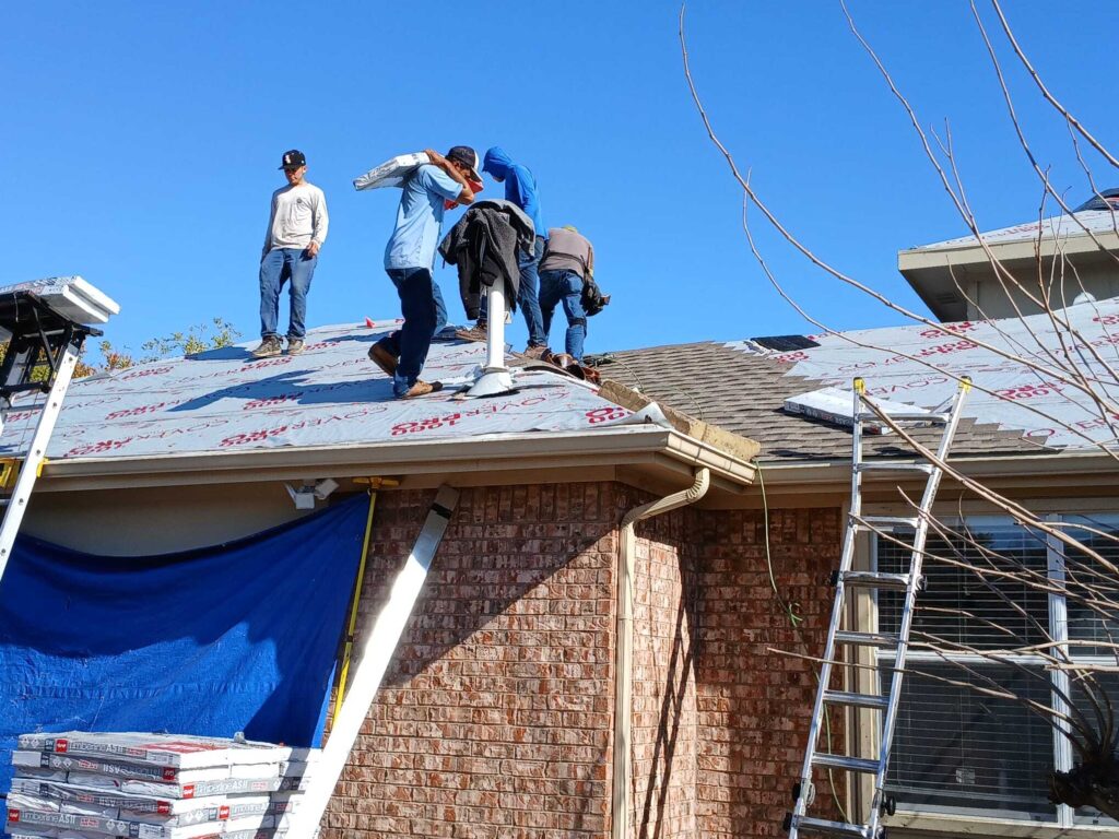 Several roofers are actively working on a residential roof, with some carrying bundles of new shingles while underlayment is already installed. Stacks of GAF Timberline ASII shingles are visible on the ground next to the house.