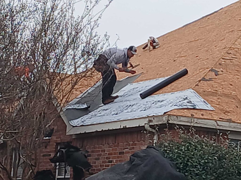 A roofer is actively installing underlayment on a residential roof, with the plywood roof deck visible in the background. The worker is wearing a cap and holding tools.