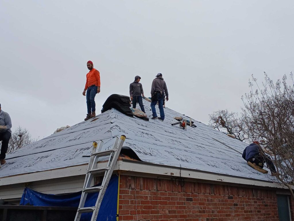 Several roofers are actively working on a residential roof, which is covered with underlayment and has bundles of new shingles ready for installation. A ladder is propped against the side of the house.