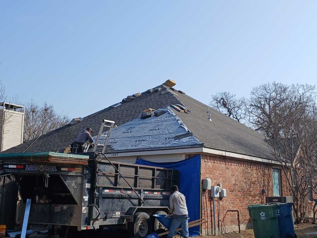 Two roofers are actively working on a residential roof, with one on a ladder near a dump trailer and another on the ground. Part of the roof has new underlayment installed, while other sections show existing asphalt shingles.