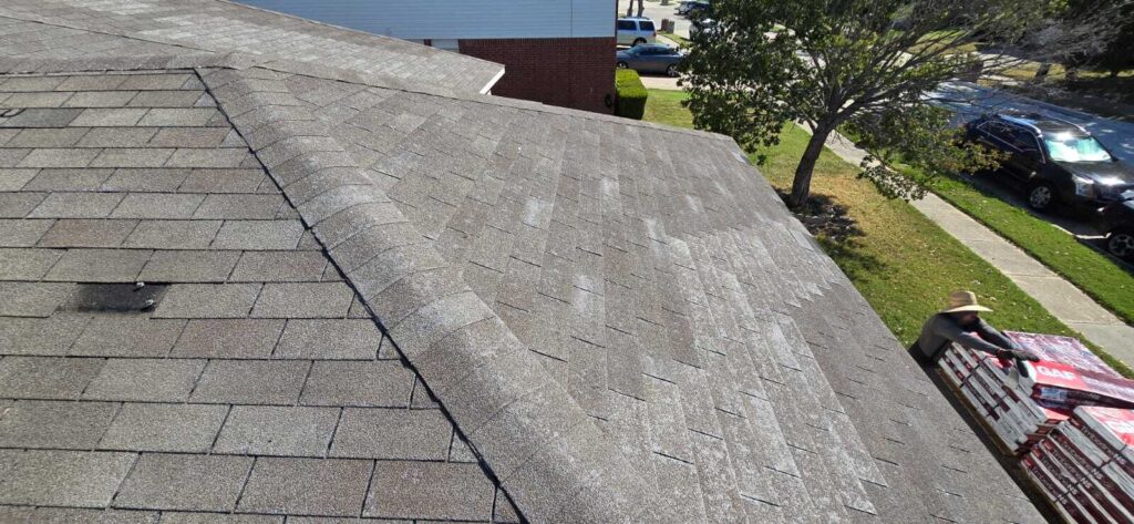 An aerial view of an asphalt shingle roof showing some wear and a missing shingle, with a worker in a straw hat on a ladder or lift next to stacks of GAF Timberline shingles. A street with parked cars and a tree is visible in the background.
