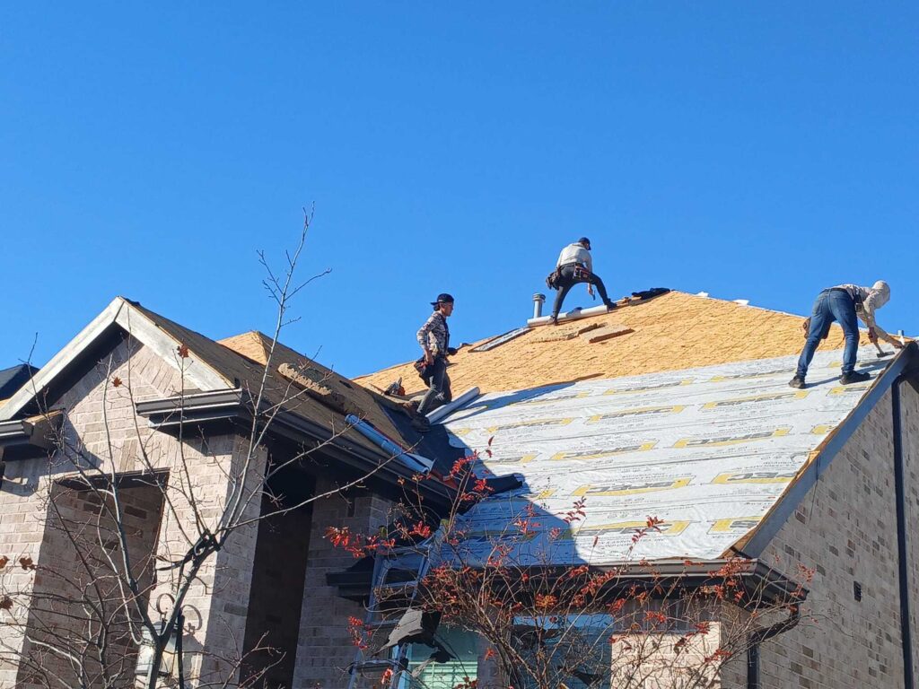 Three roofers are actively working on a residential roof, with parts of the roof showing exposed plywood decking and other sections covered with underlayment.
