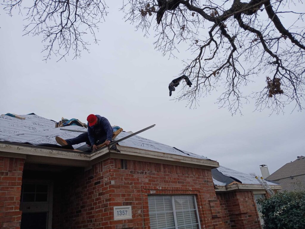 A roofer in a red cap is actively working on a residential roof, which is partially covered with underlayment. The brick house has a visible house number, and bare tree branches frame the top of the image.