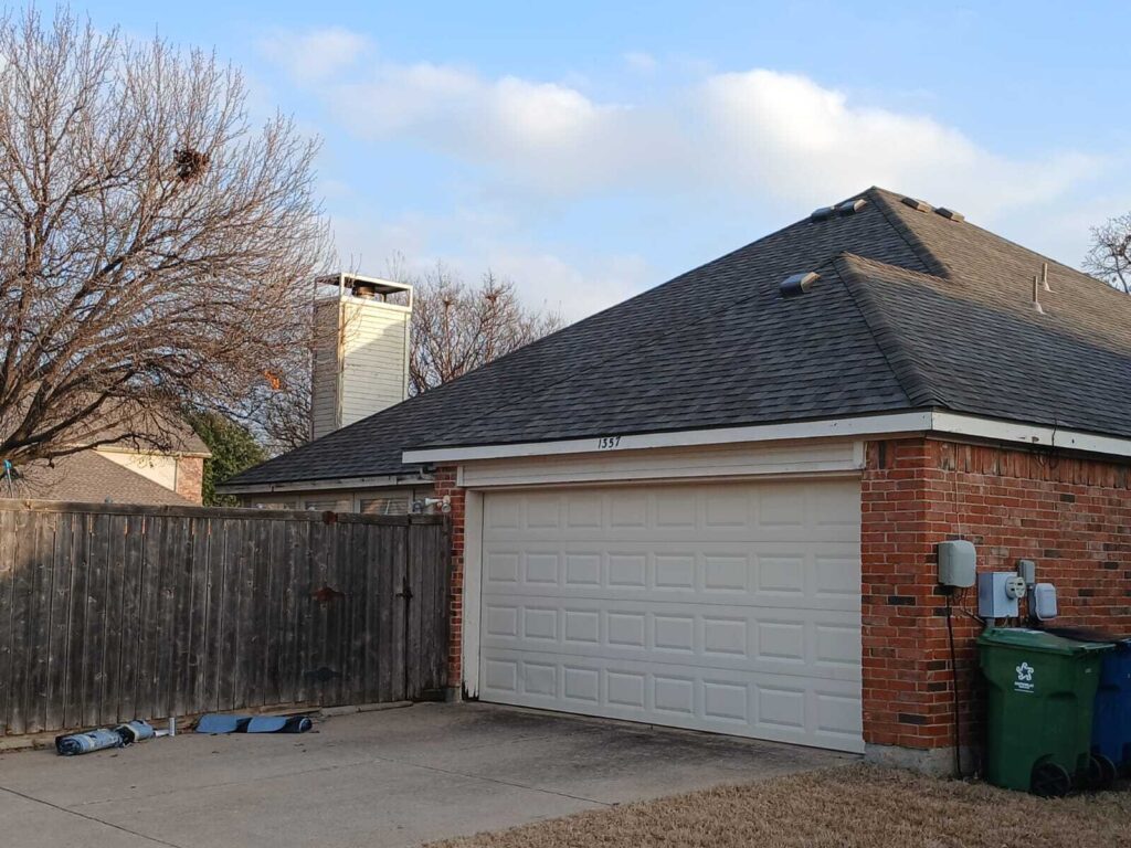 A residential house with a dark asphalt shingle roof, a white garage door, and a brick facade, viewed from the driveway. A wooden fence and bare trees are visible on the left.
