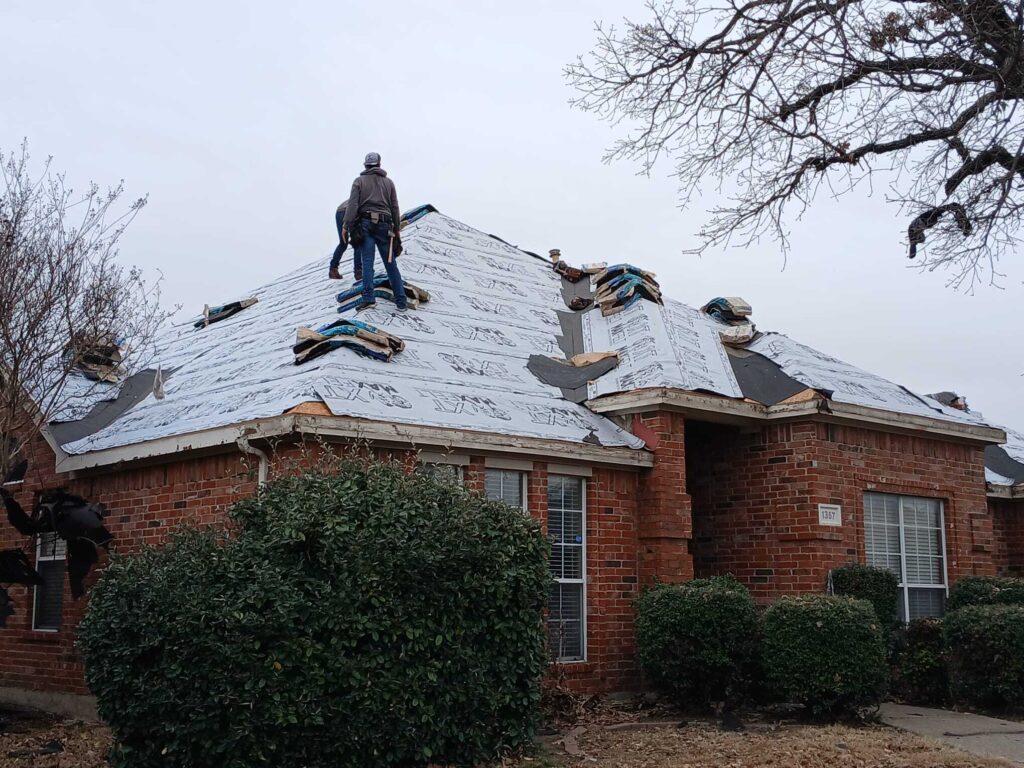 Two roofers are actively working on a residential roof, which is currently covered in underlayment. Bundles of new shingles are staged on the roof, ready for installation.