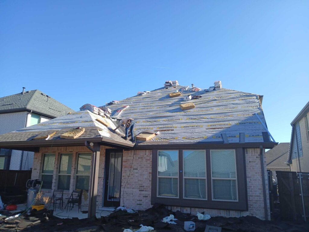 A residential roof is shown during installation, with underlayment applied and bundles of asphalt shingles stacked on the roof. One worker is visible on the roof, actively working near the eave.