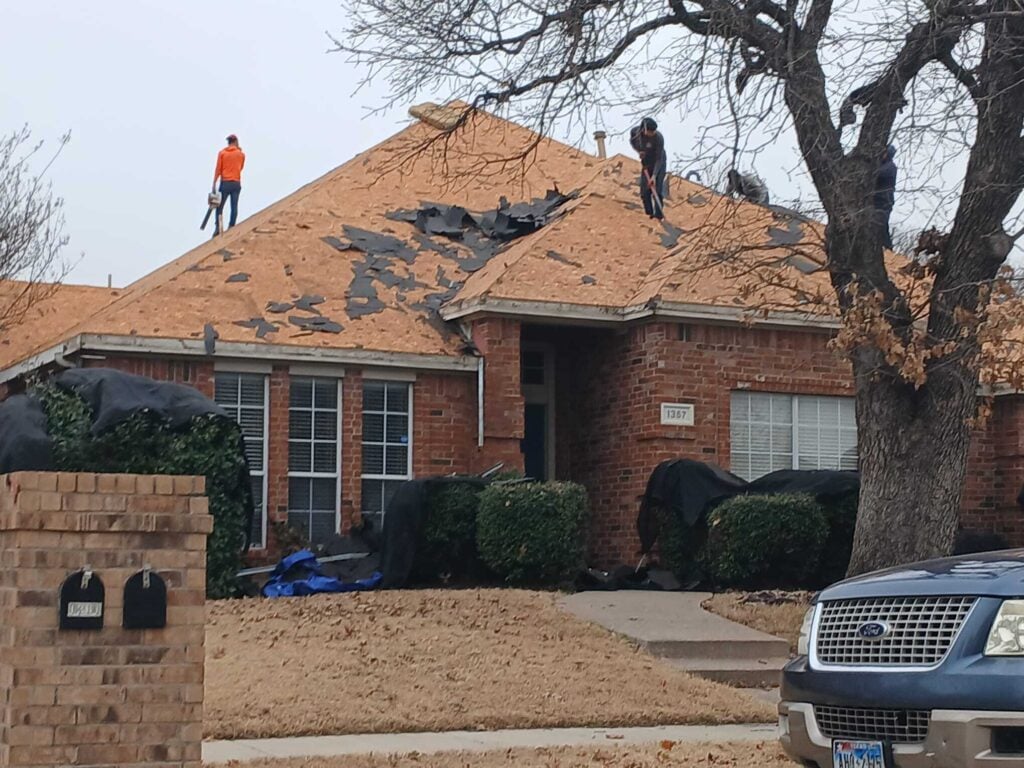 Several roofers are actively tearing off old asphalt shingles from a residential brick house under an overcast sky. Protective tarps are visible around the base of the house and over shrubs.
