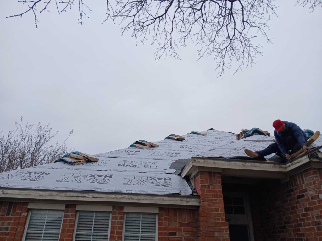 A roofer wearing a red cap is working on a residential roof covered with underlayment, with bundles of shingles visible. The house features brick siding and windows with blinds, under an overcast sky with bare tree branches overhead.